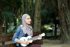 jovem ásia malaio mulher lenço de cabeça ao ar livre verde sentar em parque Banco jogar ukulele música cantar foto