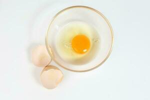 cru ovo gema e branco líquido dentro transparente vidro tigela casca de ovo branco fundo foto