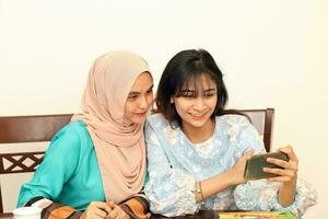 dois jovem ásia malaio muçulmano mulher vestindo lenço de cabeça às casa escritório aluna sentado às mesa telefone computador livro documento selfie auto retrato com Smartphone foto