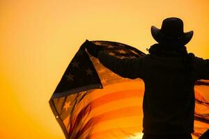 homem dentro uma vaqueiro chapéu acenando a americano bandeira às pôr do sol foto