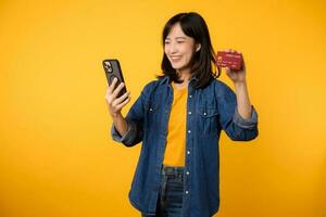 retrato lindo jovem ásia mulher feliz sorrir vestido dentro jeans Jaqueta mostrando Smartphone e crédito cartão isolar em amarelo estúdio fundo. compras conectados Smartphone inscrição conceito foto