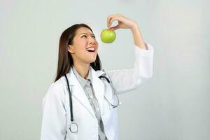jovem ásia fêmea médico vestindo avental uniforme túnica estetoscópio segurando apontando mostrando comendo saudável verde maçã foto
