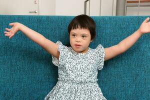 sudeste ásia pequeno pequeno menina criança sentado em azul sofá Veja feliz sorrir pose aberto braço feliz abraço cantar. ela ter baixa síndrome foto