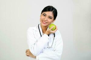 jovem ásia fêmea médico vestindo avental uniforme túnica estetoscópio segurando apontando mostrando comendo saudável verde maçã foto