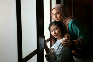 dois jovem ásia malaio muçulmano mulher vestindo lenço de cabeça baju Kurung vestir às casa dentro frente janela humor luz em face Sombrio fundo pacífico conteúdo calma tranquilo pausa cocho maravilha feliz foto