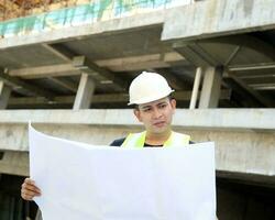 sul leste ásia jovem malaio homem vestindo branco segurança capacete amarelo colete olhando carregando projeto plano ao ar livre construção local foto