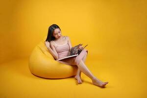 lindo jovem sul leste ásia mulher sentar em uma amarelo laranja beanbag assento cor fundo relaxar ler estude livro revista foto