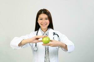 jovem ásia fêmea médico vestindo avental uniforme túnica estetoscópio segurando apontando mostrando comendo saudável verde maçã foto