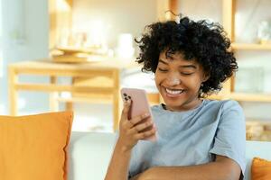 animado feliz jovem Preto africano americano mulher segurando célula telefone rindo sentindo-me alegria obtendo Móvel mensagem. muito feliz menina rindo em voz alta sentado em sofá assistindo engraçado vídeo lendo notícias. foto