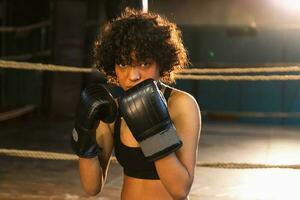 clamor independente menina poder. Bravo africano americano mulher lutador com boxe luvas olhando sério agressivo em pé em boxe anel. Forte poderoso lutador menina Treinamento socos. foto