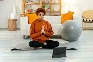 ioga atenção meditação. jovem saudável africano menina praticando ioga às lar. mulher sentado dentro lótus pose em ioga esteira olhos fechar mãos dentro oração meditando interior. menina fazendo respiração prática. foto