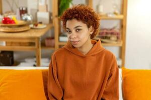 linda garota afro-americana com penteado afro sorrindo sentado no sofá em casa interior. jovem mulher africana com cabelos cacheados sorrindo. liberdade felicidade despreocupado conceito de pessoas felizes. foto