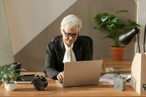 confiante à moda europeu meio envelhecido Senior mulher usando computador portátil às ambiente de trabalho. à moda Mais velho maduro anos 60 cinzento cabelos senhora empresária sentado às escritório mesa. patrão líder professor profissional trabalhador. foto