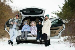 família com crianças sentar em carro suv com aberto tronco ficar de pé dentro inverno floresta. foto