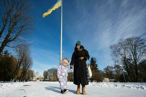 mãe e criança caminhando em uma ensolarado gelado inverno dia dentro a parque, contra fundo do uma mastro de bandeira com a ucraniano bandeira. foto