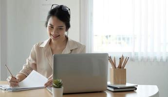 Feliz jovem empresário asiático usando computador olhando para a tela trabalhando na internet sente-se na mesa do escritório, sorrindo, funcionária profissional, digitando e-mail no laptop no local de trabalho foto