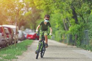 criança feliz se divertindo perto de casa com uma bicicleta em um lindo dia de primavera usando máscara de proteção para o vírus pandêmico coronavírus covid19