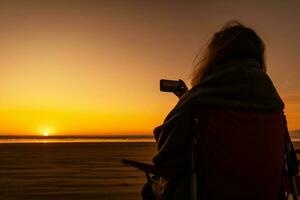 mulher coberto de caloroso cobertor levando vídeo do uma cênico de praia pôr do sol foto