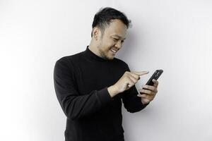 animado ásia homem vestindo Preto camisa sorridente enquanto segurando dele telefone, isolado de branco fundo foto