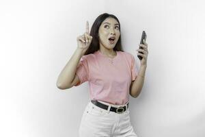 uma retrato do uma chocado ásia mulher vestindo Rosa camiseta, segurando dela telefone, e apontando cópia de espaço em topo do dela, isolado de branco fundo foto