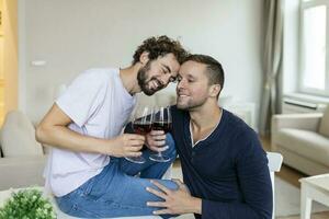 lgbtq casal abraçando cada de outros e bebendo vinho dentro de casa. dois romântico jovem masculino amantes olhando às cada de outros enquanto sentado juntos dentro seus vivo sala. jovem gay casal ser romântico às lar. foto