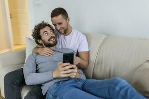 fofa jovem gay casal vídeo chamando seus amigos dentro seus vivo quarto às lar. dois masculino amantes sorridente alegremente enquanto cumprimento seus amigos em uma Smartphone. jovem gay casal sentado junto. foto