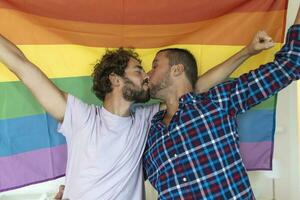 dois jovem gay amantes se beijando cada de outros carinhosamente. dois jovem masculino amantes em pé juntos contra uma orgulho bandeira. afetuoso jovem gay casal partilha uma romântico momento junto. foto