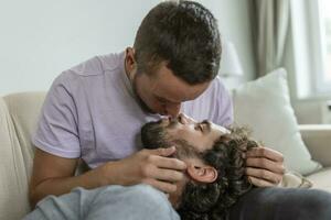 adorável gay casal se beijando enquanto sentado dentro seus vivo quarto às lar. dois romântico jovem masculino amantes tendo Diversão dentro de casa. jovem gay casal vivo junto. foto