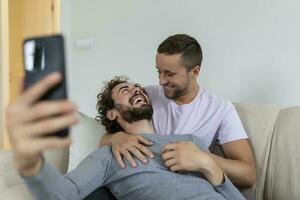 fofa jovem gay casal vídeo chamando seus amigos dentro seus vivo quarto às lar. dois masculino amantes sorridente alegremente enquanto cumprimento seus amigos em uma Smartphone. jovem gay casal sentado junto. foto