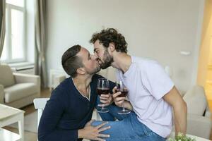 lgbtq casal abraçando cada de outros e bebendo vinho dentro de casa. dois romântico jovem masculino amantes olhando às cada de outros enquanto sentado juntos dentro seus vivo sala. jovem gay casal ser romântico às lar. foto