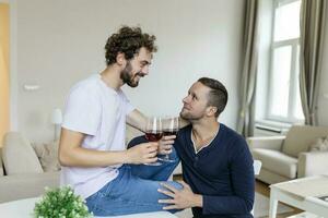 lgbtq casal abraçando cada de outros e bebendo vinho dentro de casa. dois romântico jovem masculino amantes olhando às cada de outros enquanto sentado juntos dentro seus vivo sala. jovem gay casal ser romântico às lar. foto