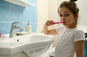 fechar-se adorável bebê menina vestido dentro pijamas, levando Cuidado do dela dentes, escovar dentes com pasta de dentes e escova de dente foto