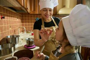 sorridente alegre mãe dando uma solta do chocolate para dela filha enquanto cozinhando doce sobremesa juntos dentro a casa cozinha foto