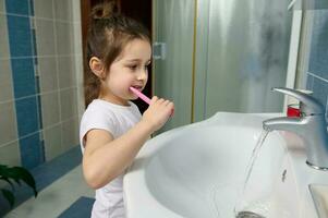 lindo pré-escolar pequeno menina escovar dentes, em pé de Pia dentro a casa banheiro. dental Cuidado e oral higiene. foto