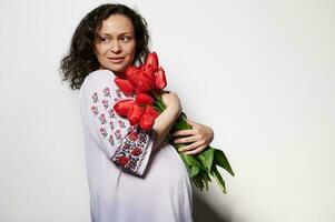 encantador grávida mulher, vestindo bordado camisa com ucraniano étnico ornamento, posando com uma ramalhete do vermelho tulipas foto