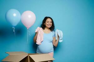 feliz grávida mulher mostrando uma azul e Rosa recém-nascido maiô, expressando espanto às gênero revelar Festa. bebê chuveiro foto