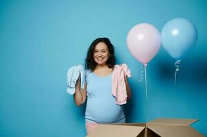 lindo grávida morena mulher sorrisos, olhando às Câmera, detém recém-nascido bebê macacões sobre isolado azul fundo foto