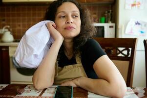 pensativo jovem mulher dona de casa, padeiro pasteleiro dentro chef avental, sentado às mesa dentro casa cozinha, olhando longe foto