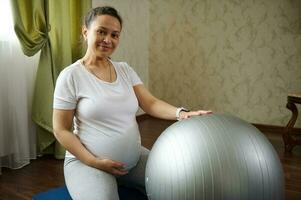 autêntico grávida mulher sorrisos às Câmera, toques dela barriga, sentado em uma ioga esteira enquanto enquanto exercício com em forma bola foto