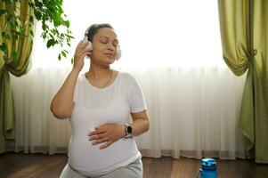 grávida mulher escuta para música em fones de ouvido, suavemente carícias dela barriga pensando sobre dela bebê, desfrutando dela gravidez foto