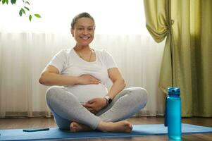 feliz adulto grávida mulher suavemente acariciando dela barriga, sentado dentro lótus pose em uma esteira durante gravidez ioga prática foto