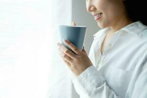 fechar-se imagem do uma lindo jovem ásia mulher cheirando e bebendo café. ela sentado perto a janela dentro a manhã. foto