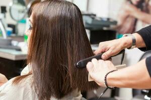 estilista usando plano ferro em cabelo do fêmea cliente sentado dentro salão cadeira foto