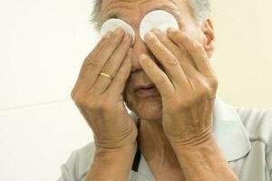 Senior homem dentro a banheiro usando algodão almofadas para olho alívio foto