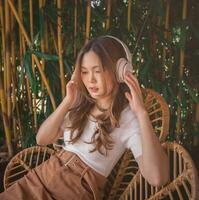 relaxado ásia mulher com sem fio fones de ouvido, ouvindo para meditação música e sorridente com prazer. alegre jovem menina desgasta branco camiseta sentado em cadeira dentro acolhedor botânico jardim. estilo de vida. foto