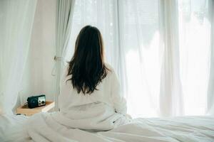 jovem asiática de pijama branco acordando em sua cama sentada perto da grande janela totalmente descansada e abrindo as cortinas pela manhã para tomar ar fresco e sol. jovem feliz cumprimenta o novo dia. foto