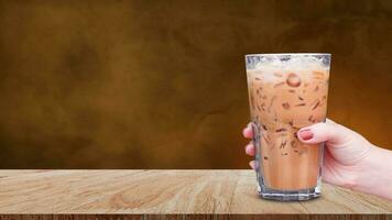 mão segurando a vidro gelado café em de madeira mesa em abstrato Sombrio fundo, gelo café com leite café foto