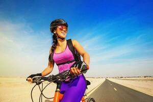 jovem sorridente caucasiano mulher equitação bicicleta em uma estrada dentro deserto foto