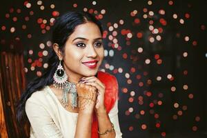 jovem lindo indiano mulher vestindo joalheria e festivo étnico roupas sentado e esperando sobre bokeh fundo foto