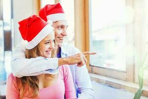 uma casal vestindo Natal santa vermelho chapéus abraçando. homem apontando às a janela foto